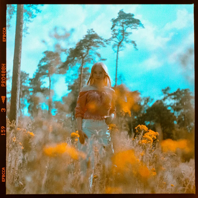 Analoges Portrait einer Frau in einem Feld mit gelben Wildblumen und Kiefern im Hintergrund.