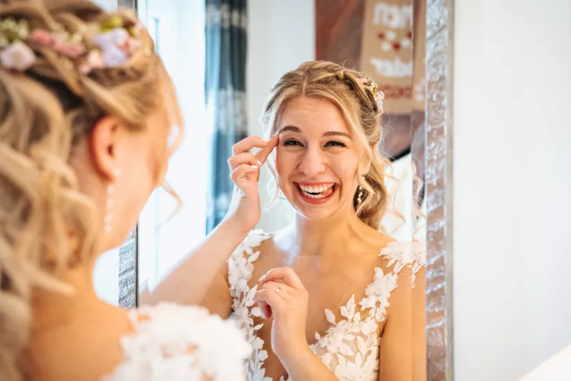text: "Lachende Braut mit Blumen im Haar beim Blick in den Spiegel