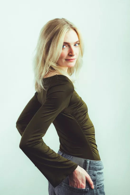 Portrait einer blonden Frau im olivgrünen Oberteil und Jeans vor hellem Hintergrund